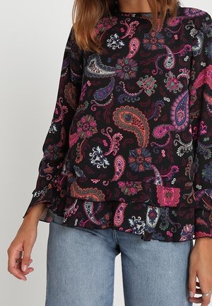 Donna che indossa una maglia nera a maniche lunghe con motivi paisley e floreali colorati rosa, viola e arancione, abbinata a jeans azzurri chiari.