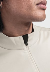 Helles beiges Sportjacke mit hohem Kragen, ausgestattet mit einem schwarzen Reißverschluss und strukturiertem Stoff. Nahaufnahme des Halsausschnitts.