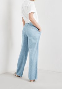 Persoon staat met handen in de zakken van lichtblauwe wijde broek, en draagt een witte kortarmsgemouwen blouse en witte platte schoenen.