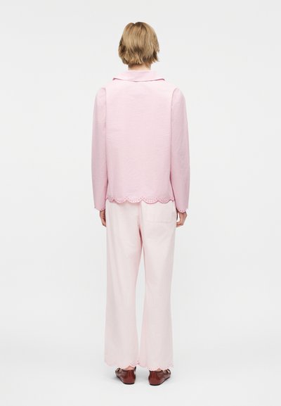 Άτομο που στέκεται με την πλάτη γυρισμένη, φορώντας ανοιχτό ροζ μακρυμάνικη μπλούζα με κυματιστό τελείωμα και αντίστοιχο παντελόνι φαρδιάς γραμμής, με σκούρα καφέ παπούτσια.