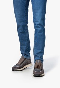 Piernas vestidas con jeans azules y zapatillas gris y beige con cordones, de pie sobre una superficie blanca.