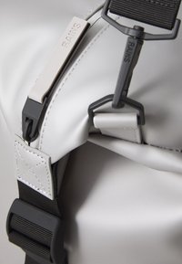 Primo piano di una borsa elegante di colore grigio chiaro, dotata di una fibbia e una tracolla, con l'etichetta del marchio "RAINS" visibile sul tirante della cerniera.
