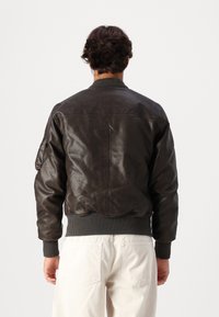 Jacket bomber de couro castanho com punhos e barra canelados, apresentando um bolso lateral e uma textura suave. O design é justo e clássico.