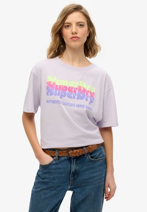 T-shirt oversize lavande avec un graphique "Superdry" multicolore. Col rond, manches courtes, associé à un jean bleu et une ceinture tressée marron.
