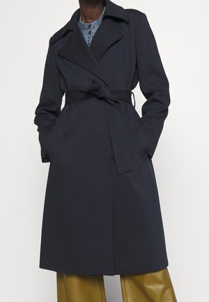 Hombre con un abrigo tipo trench azul marino con cinturón sobre una camisa azul claro y pantalones anchos color mostaza, con las manos en los bolsillos del abrigo.