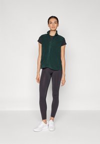 Chaleco de forro polar verde con cremallera y cuello alto, usado sobre una camiseta negra, combinado con leggings negros y zapatillas blancas. Diseño simple y ligero.
