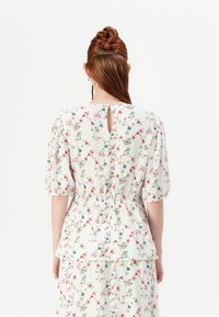 Blusa floreale con maniche a palloncino corte, vita elastica e schiena a chiave. Tessuto bianco con piccoli fiori rossi e blu. Texture morbida e leggera.