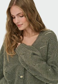 Cardigan verde a maglia con scollatura a V, vestibilità ampia e bottone decorativo. Presenta un tessuto strutturato e motivi a occhiello sulle maniche.