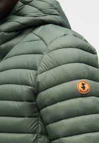 Oliivinvihreä paksupuntainen takki, jossa vaakasuoria tikkauksia ja pyöreä oranssi kangasmerkki vasemmassa olkapäässä, jossa on ankkalogo.