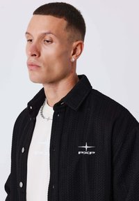 Schwarze gewobene Hemdjacke mit einem strukturierten Muster, Druckknöpfen und einem Logo auf der Brust. Getragen über einem hellen T-Shirt.