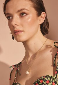 Zelta ķēdes kaklarota ar apaļu pērlīšu piekariņu un mazu akcenta dārgakmeni. Modelis valkā ziedu raksta apģērbu ar sasietām siksnām.