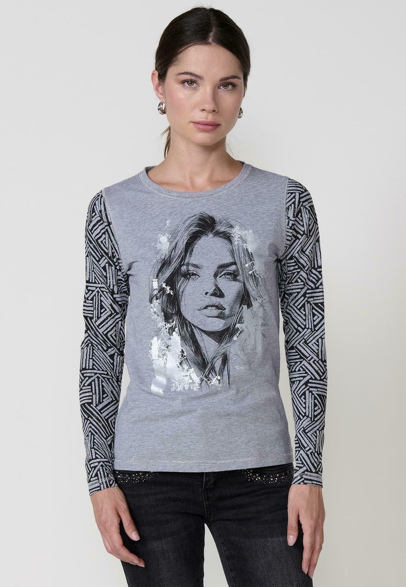 La camiseta de manga larga gris presenta un gráfico central plateado de una cara, con mangas negras estampadas y un escote redondo.
