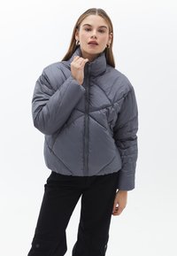 Chaqueta de abrigo gris con un alto cuello, diseño de acolchado en diamante y cremallera frontal. Lleva puesta sobre pantalones negros con bolsillos laterales.