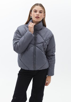 Chaqueta de abrigo gris con un alto cuello, diseño de acolchado en diamante y cremallera frontal. Lleva puesta sobre pantalones negros con bolsillos laterales.