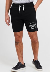 Zwarte katoenen shorts met een witte trekkoord en een logo in schrijfstijl. Draagt zwarte sneakers en toont een tatoeage op het been.