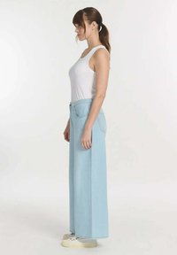 Lichtblauwe wijde denimbroek met hoge taille, voorzien van voorzakken en een subtiele stikseldetail, gedragen met een witte tanktop.