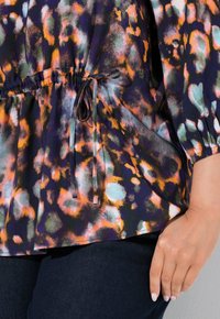 Bunt gemusterte Bluse mit einem gerafften, elastischen Bund und einem Bindeband, kurzen Ärmeln und einem fließenden Design. Der Stoff zeigt lebendige, abstrakte Muster.