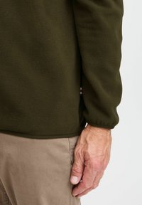 Pull en polaire vert olive avec une texture douce et des poignets élastiques, associé à un pantalon beige. Présente une petite étiquette logo multicolore.