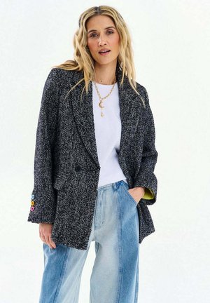 Blondynka w ciemnoszarej marynarce w jodełkę z kolorowymi guzikami, białej koszuli, naszyjniku w kształcie półksiężyca i dżinsach z szerokimi nogawkami w dwóch odcieniach.