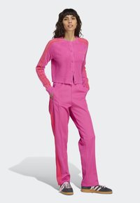 Woman standing with hands in pockets wearing bright pink fuzzy cardigan and matching track pants with red stripes, and black sneakers with pink laces.