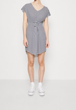 Kobieta ubrana w granatowo-białą sukienkę w paski z krótkim rękawem i sznurkiem w talii oraz białe platformowe trampki, stojąca na tle jednolitej białej ściany.