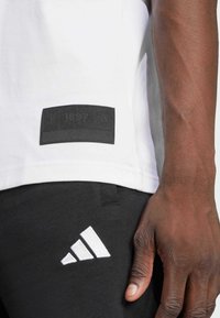 White cotton t-shirt with a black fabric patch featuring embossed logo, paired with black pants displaying a white three-stripe logo.