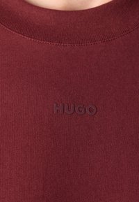Burgundi pulóver bordás nyakkivágással. Diszkrét, azonos színű, dombornyomott "HUGO" logóval rendelkezik. Sima textúrájú és testhezálló siluettel.
