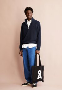 Sudadera azul marino con cuello de cremallera sobre una camisa blanca, combinada con unos vaqueros anchos azules. Bolso de mano negro con un diseño gráfico blanco; zapatos negros.