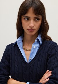 Jersey de punto trenzado en azul marino, superpuesto a una camisa de rayas en azul claro, con escote en V, acompañado de un collar de cadena dorada y tela texturizada.
