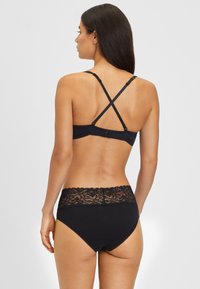 Schwarzes Dessous-Set mit einem Bralette, das verstellbare Kreuzgurte hat, und hochgeschnittenen Slip mit einem wellenförmigen Spitzenbund.