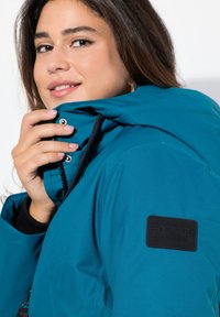 Teal isolerad jacka med huva, zippered framtill och ett svart varumärkespatch på ärmen. Slät textur med en avslappnad passform.