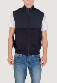 Gilet blu navy con zip e colletto alto, realizzato in un materiale liscio, con un design semplice e due tasche laterali. Indossato sopra una maglietta bianca.