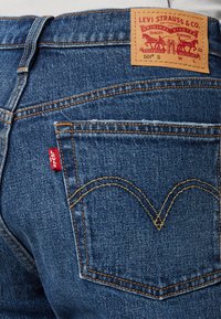 Jeans de mezclilla azul que presentan un distintivo parche de cuero marrón con la marca, costuras naranjas y una pequeña etiqueta roja en el bolsillo trasero.