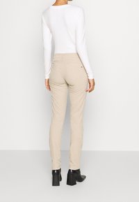 Pantaloni beige con una vestibilità slim, realizzati in un materiale morbido. Presentano tasche posteriori e dettagli di cucitura. Indossati con un top bianco a maniche lunghe attillato.