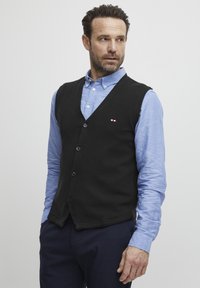 Svart ullväst med V-halsdesign, prydd med fem knappar och en liten logotypdetalj. Bärs över en ljusblå skjorta med knappar.