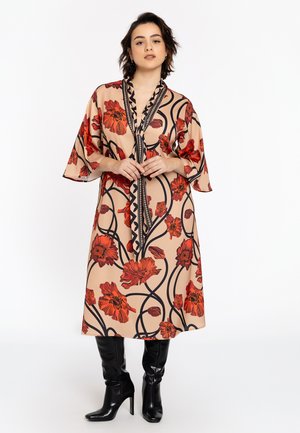 Vloeiende beige jurk met rode bloemenpatronen, zwarte omlijning en een diepe V-hals. Heeft brede mouwen en een getailleerd ontwerp. Gecombineerd met zwarte kniehoogte laarzen.