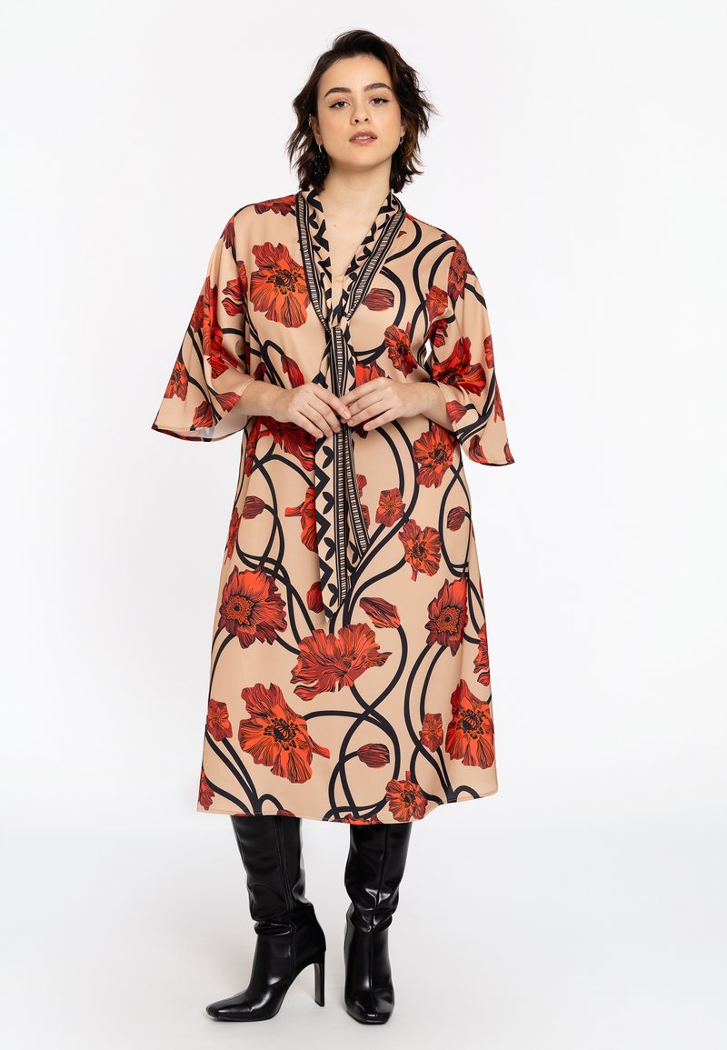 Vloeiende beige jurk met rode bloemenpatronen, zwarte omlijning en een diepe V-hals. Heeft brede mouwen en een getailleerd ontwerp. Gecombineerd met zwarte kniehoogte laarzen.