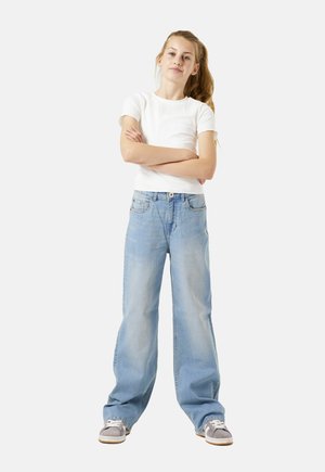 Tienermeisje met lang haar in een paardenstaart staat met de armen gekruist, gekleed in een wit T-shirt met korte mouwen, brede lichtblauwe jeans en grijze sneakers.