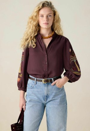 Dunkelrotes Hemd mit Knöpfen und Ballonärmeln, verziert mit mehrfarbiger Stickerei, kombiniert mit hellen Jeans und braunem Gürtel.