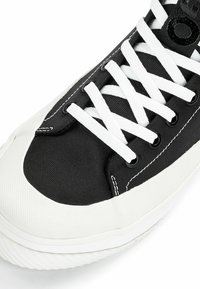 Schwarze Leinenschuhe mit weißen Schnürsenkeln, einer Gummizipfelferse und weißer Sohle. Schwarze Ösen und Nähte betonen das Design.