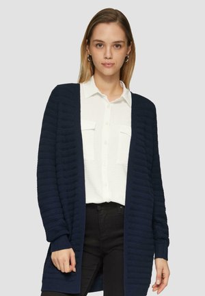 Gilet en mailles bleu marine avec un design texturé, à l'ouverture sur le devant et à manches longues, porté par-dessus une chemise blanche à boutons et un pantalon noir.