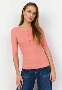 Ribbed, short-sleeve, pink top with a rounded neckline and front buttons. Paired with blue jeans, featuring standard pockets and stitching.