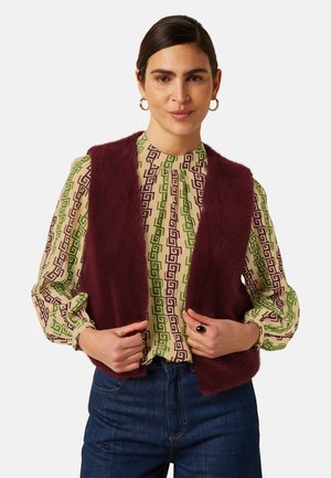Vrouw die een bordeauxrode pluizige bodywarmer draagt over een blouse met lange mouwen en patroon en blauwe spijkerbroek, die de randen van de bodywarmer met beide handen vasthoudt.