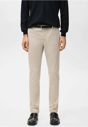 Mężczyzna ubrany w beżowe spodnie typu slim fit, czarny pasek, czarny sweter oraz czarne skórzane loafersy, stojący na jednolitym tle.