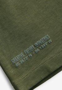 Olivgrön bomulls t-shirt med ribbad textur, med blå tryckt text: "SKAPA FRAMTIDA INDUSTRIER 40.2672° N - 86.1349° W."