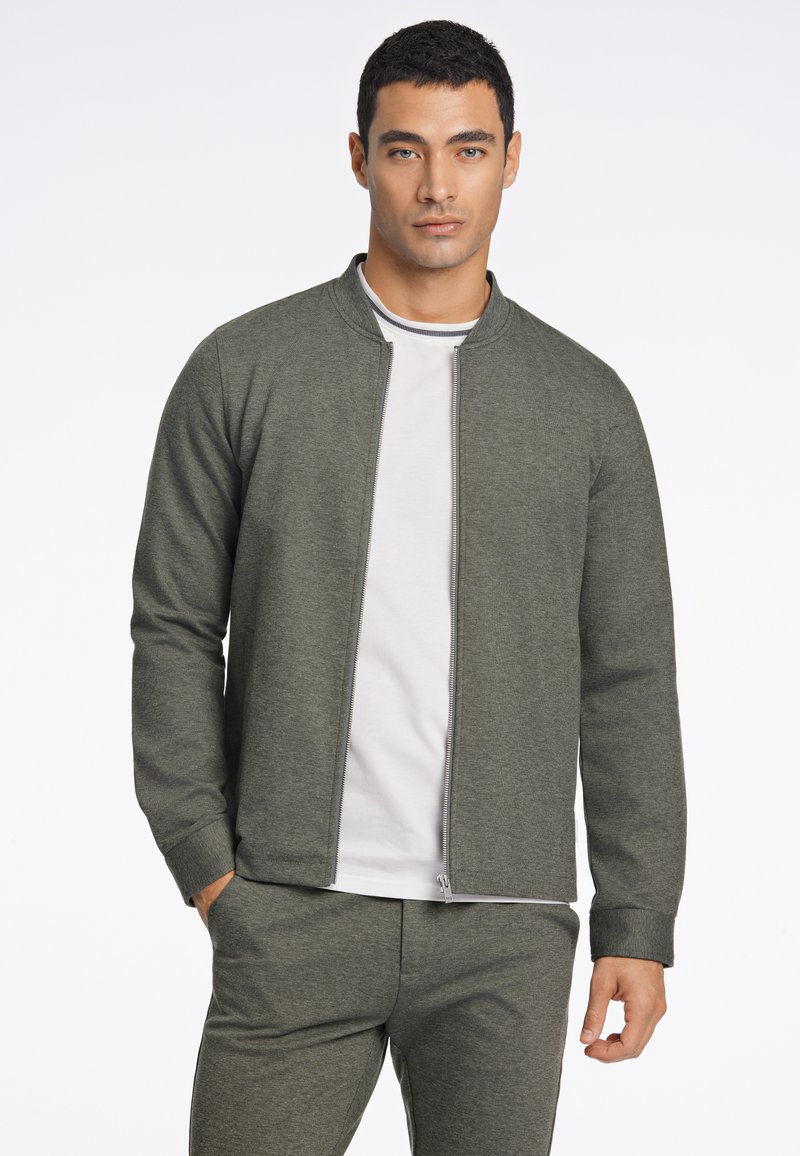Chaqueta de cremallera verde oscuro en un material texturizado con cuello alzado, combinada con una camiseta blanca de cuello redondo debajo.