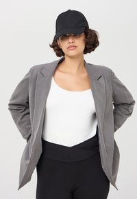 Blazer grigio con un design strutturato, indossato sopra una maglietta bianca; abbinato a pantaloni neri a vita alta e un cappellino da baseball nero.