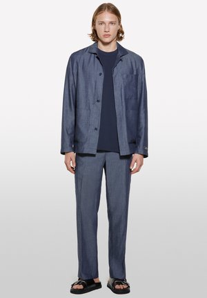 Navy textured shirt jacket and matching trousers in a lightweight fabric, featuring a pocket on the left chest and button closures. Black sandals.