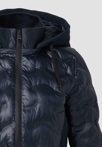 Mornarska puhovka z žakardasto teksturo, visoko ovratnico in kapuco. Ima črn zadrgo in dodatke s šnuro, ki omogočajo prilagojeno prileganje.