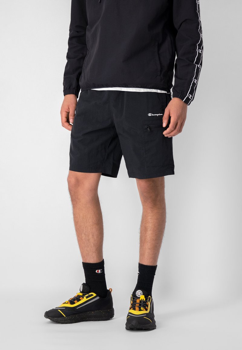 Pantaloni corti neri realizzati in tessuto leggero, con una tasca zip, cintura elastica e logo Champion. Abbinati a sneaker nere e gialle.